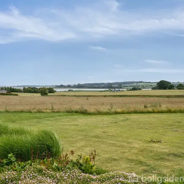 Et græsareal breder sig foran et åbent landskab med marker og enkelte bygninger i baggrunden under en blå himmel med lette skyer.