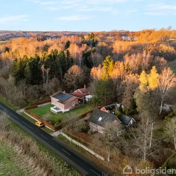 Et hus står ved siden af en snoet vej, omgivet af træer med gule og brune blade. Landskabet er landligt og bakket med en klar himmel i baggrunden.