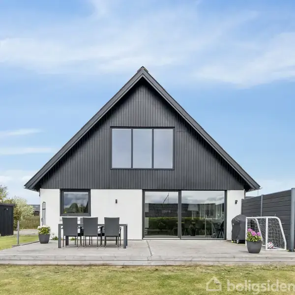 En moderne bungalow med sort gavl og store vinduer står på en grøn græsplæne. En terrassemøblement står på terrassen, mens en lille fodboldmål ses til højre.