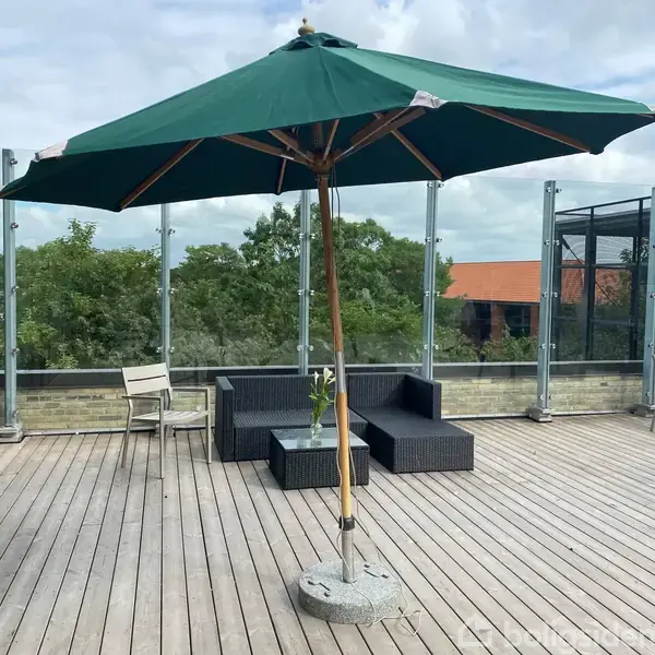 En stor grøn parasol står på en træterrasse omgivet af havemøbler. I baggrunden ses et glasrækværk og træer.