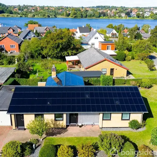 En bungalow med solpaneler på taget står i en velplejet have. Huset er omgivet af andre boliger og ligger nær en sø, set i baggrunden med en grøn bakketop.