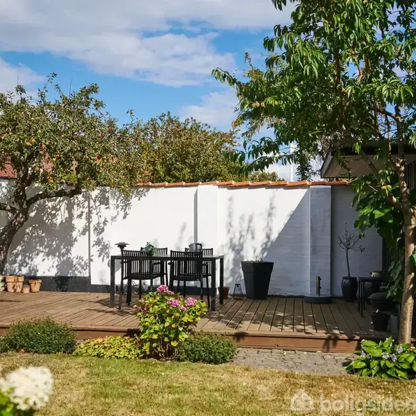 En terrasse har et bord og fire stole. Omkring terrassen ses en have med græsplæne, blomsterbede og træer. En hvid mur løber langs baggrunden under en blå himmel med skyer.