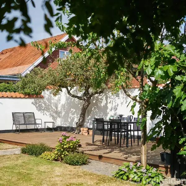 En træterrasse med havemøbler, omgivet af en have med et træ og blomsterkrukker. I baggrunden ses et hvidt hus med rødt tegltag.