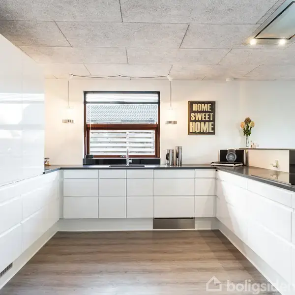 Et moderne, lyst køkken med hvide skabe omgiver en central arbejdsflade. På væggen hænger et skilt med teksten "HOME SWEET HOME". Et vindue og blomster tilføjer naturligt lys og farve.
