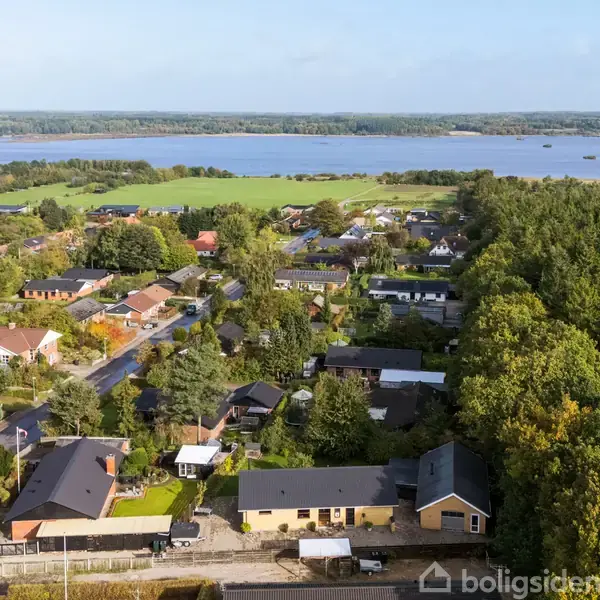 Bebyggelse med huse, der står tæt sammen, omgivet af tæt skov og åbne marker. En flod eller sø i baggrunden skaber en naturskøn udsigt over landskabet.