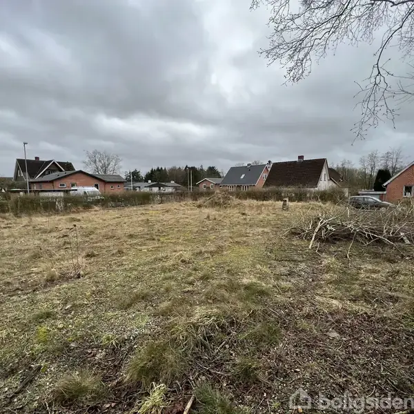 En åben græsmark ligger i forgrunden uden handling, mens der i baggrunden er huse med røde tage under en overskyet himmel. En bil er synlig til højre.