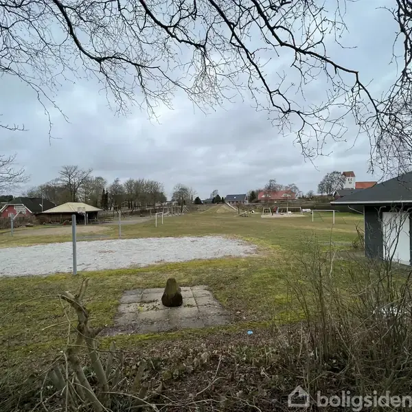 En flad græsplæne med en sandbane ligger omgivet af buske og træer. I baggrunden ses få bygninger under en overskyet himmel.