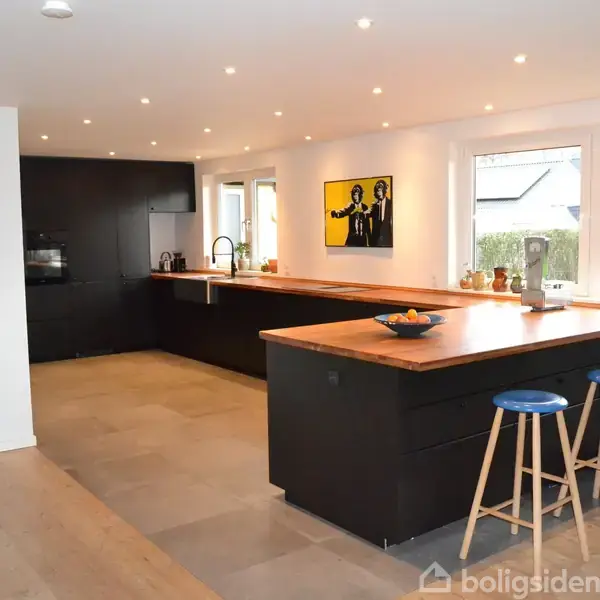 Et moderne køkken med mørke skabe og lys træbordplade. Tre blå barstole står ved øen. Billedkunst pryder væggen, omkranset af store vinduer med udsigt til have.