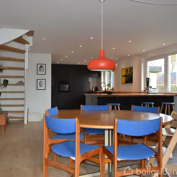 En træspisebord med blå stole står centralt. En orange pendellampe hænger over bordet. Til venstre er en trappe med planter ved siden af. Køkkenet ses i baggrunden.