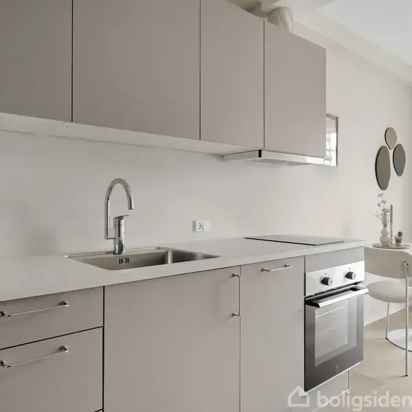 Køkken med grå skabe og hvid bordplade; en indbygget ovn under kogeplader; vask med hane. I baggrunden står et rundt spisebord med to stole ved et vindue.