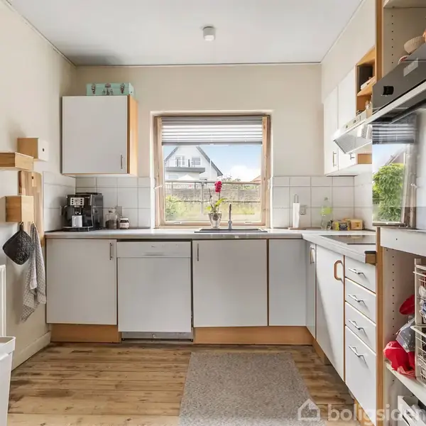 Køkken med hvidt inventar, træbordplade og trægulv. Kaffemaskine og planter på bordet. Vindue med udsigt til have og hus. Åben hylde og ovn til højre.