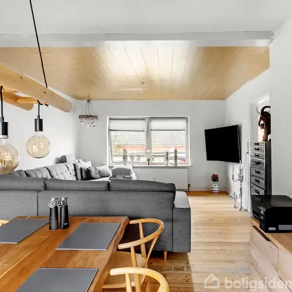 Gråt sofaarrangement står i centrum, omgivet af en spisestue med træbord og stole til venstre. Væghængt fladskærms-TV og kunstværker pryder rummet med lyse trædetaljer. Lysindfald fra vindue.