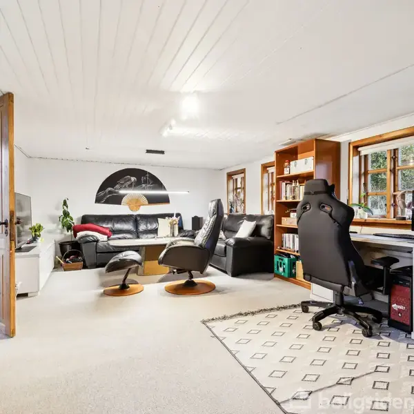 Et møbleret værelse med en kontorstol foran et skrivebord med computer. Sofaer og stole danner en siddegruppe bagved. Hylder fyldt med bøger ved vinduerne giver en hyggelig stemning.