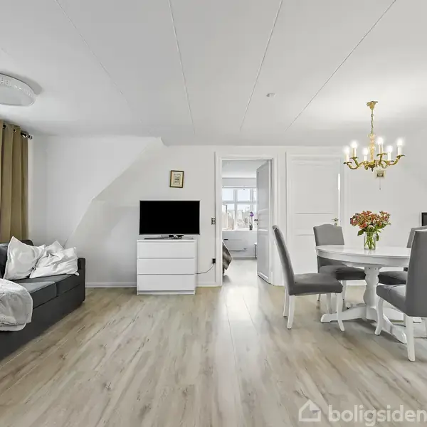 En stue med en sort sofa til venstre, placeret foran store vinduer med gardiner. Et tv står foran sofaen. Spisebord omgivet af fire grå stole og en lysekrone hænger over det. I baggrunden ses et køkken og en åbning til et andet rum.