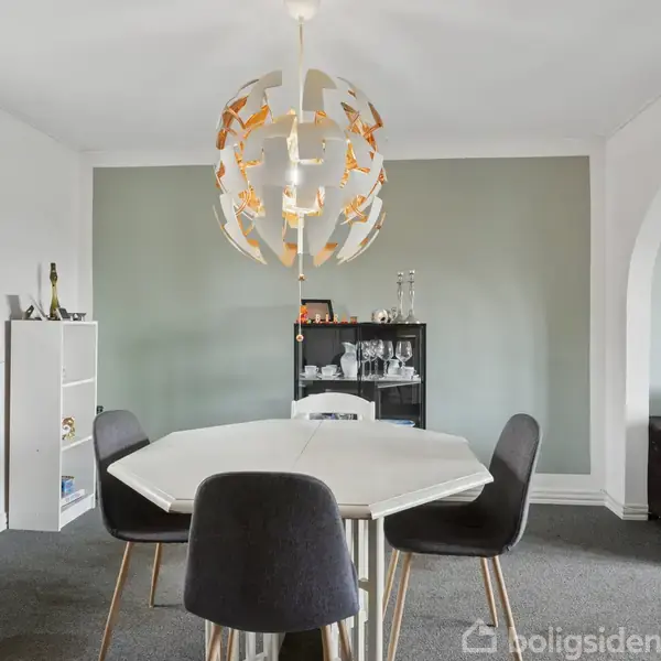 Rundt spisebord med fire sorte stole står i et lyst rum. Over bordet hænger en dekorativ lampe. Bagved ses en reol med glas og flasker. Til højre er en grå sofa.