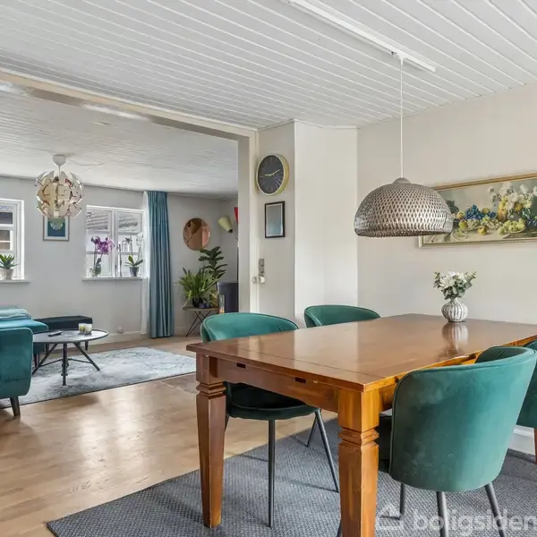 En spisebord i træ er omgivet af turkisblå stole i en stue. En stor, rund lampe hænger over bordet. Baggrunden viser en opholdsstue med sofaer og planter.