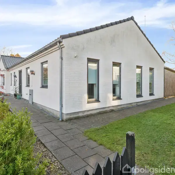 Hvidt hus med sorte tagsten, stående på en velholdt plæne med belagt sti bredved. Omgivet af træer og buske, der skaber en rolig atmosfære.