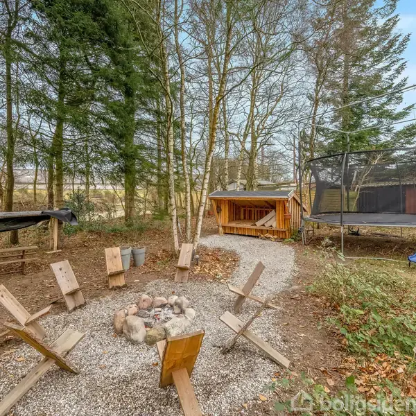 En bålplads med seks træstole rundt om en stencirkel. I baggrunden er en trampolin, et skur og et bord dækket med en presenning. Omkring er der træer.