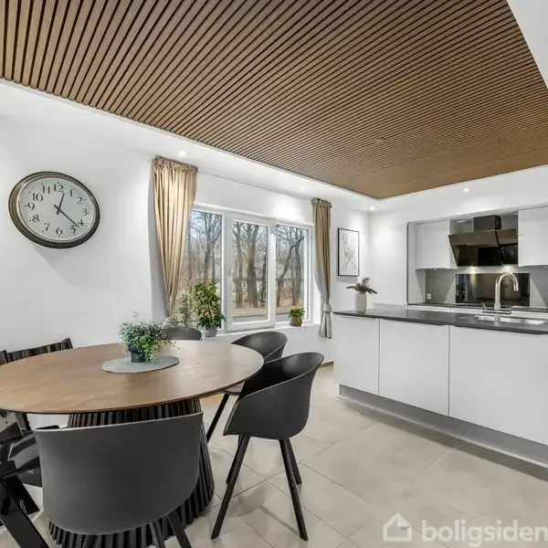 Rundt spisebord med sorte stole placeret i et moderne køkken med hvide skabe, spejle og indbyggede apparater. Store vinduer med udsigt til træer. Vægur hænger på væggen.