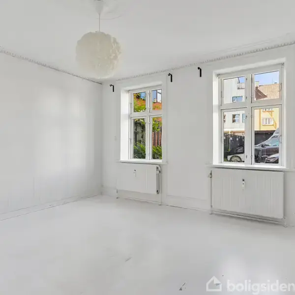 Et tomt, hvidt værelse med to vinduer, der slipper lys ind, og en hængende lampe i loftet. Værelserne viser en gade og en bil udenfor.