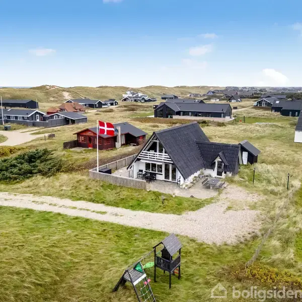 Et sort stråtækt hus står i et græsklædt område med en dansk flagstang. Omgivet af flere fritliggende huse under en blå himmel med spredte skyer.