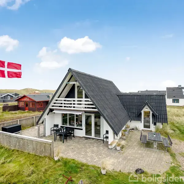 Et sommerhus med spidstag står på en græsplæne under en blå himmel. Et dansk flag vajer ved siden af en terrasse med spisebord og stole.