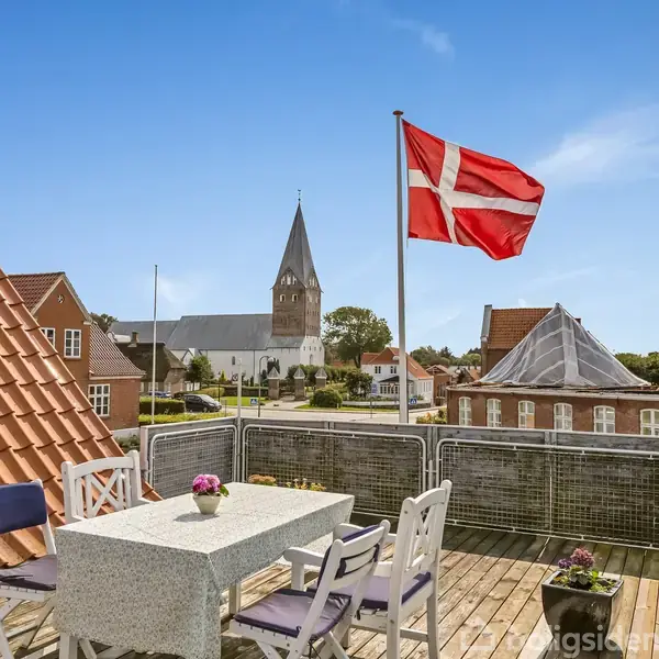 Bord med stole står på en balkon i solen med udsigt over en landsby. En Dannebrog-flag blafrer i vinden. I baggrunden ses en kirke og flere bygninger.