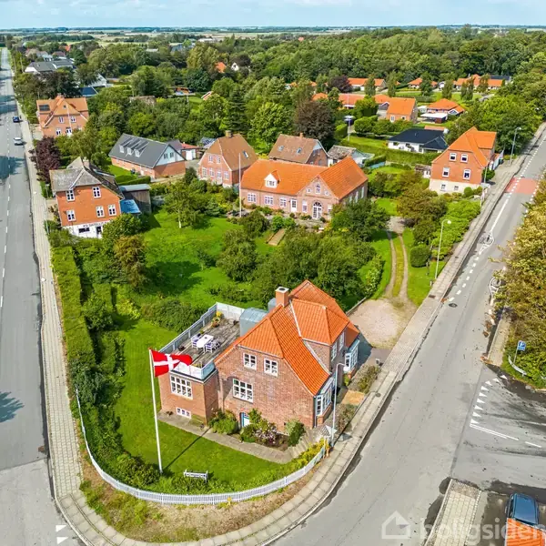 Et lyst rum med et rundt spisebord og stole i forgrunden, placeret foran store vinduer, der viser en grøn udsigt. En reol og kommode står ved siden af en hoveddør.