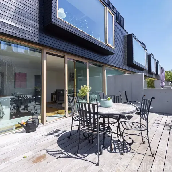Et rundt bord med stole står på en træterrasse. Terrassen ligger foran en moderne bygning med store glasvinduer, der reflekterer en klar himmel. Planter og dekorationer ses omkring.