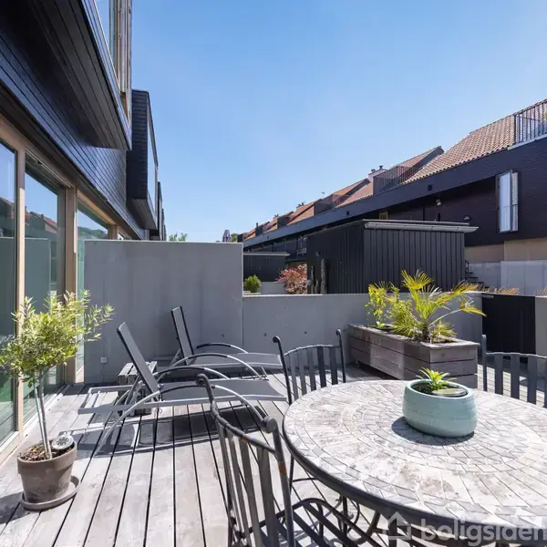 En udendørs terrasse rummer en rund marmorbord med sorte stole, omgivet af planter i potter. Solsenge står mod en moderne boligblok med store glasdøre. Skyfri himmel.