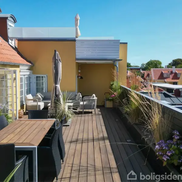 En tagterrasse med træmøbler og plantekrukker under lyseblå himmel. Omgivet af gul murbygning og udsigt over røde hustage. Store parasoller står langs spisebordet.