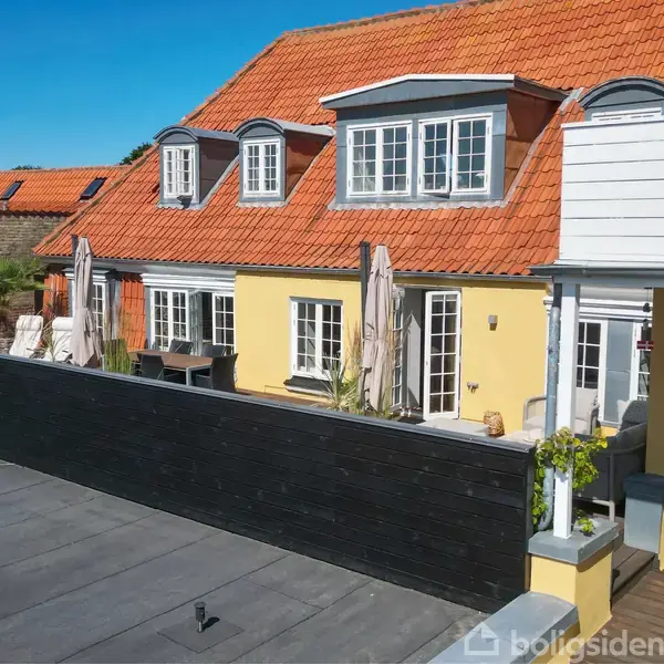Terrasse med bord og stole i forgrunden, flere planter omkring. I baggrunden ses gulmalede bygninger med rødt tag. Klar blå himmel danner en solrig og afslappet atmosfære.