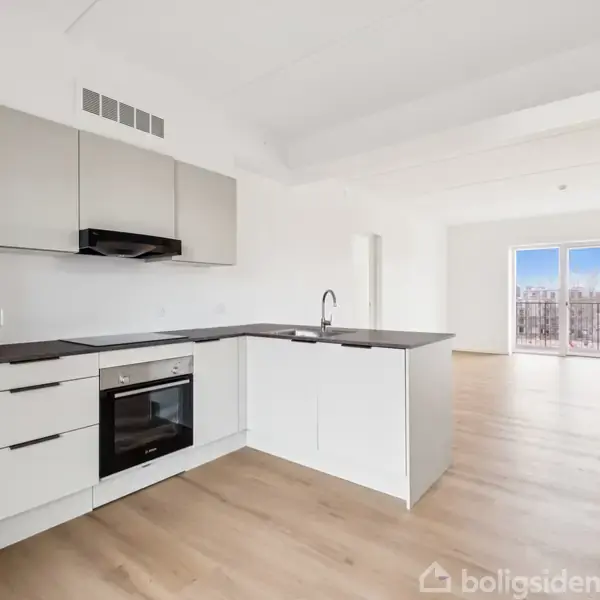 Moderne hvidt køkken med sort bordplade og indbygget ovn. Køkkenet er i et lyst rum med trægulve, der fører til en stue med vinduer og udsigt udenfor.