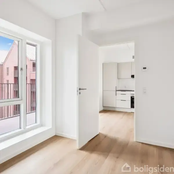 Et lyst, tomt rum med trægulv og et stort vindue til venstre, viser en del af en bygningsfacade udenfor. En åben dør fører ind til et køkken.