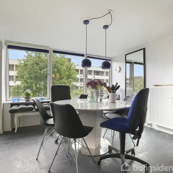 Rundt bord med sorte stole omkranset af store vinduer. Bordet har blomster og lamper hænger over det. Baggrunden viser en lys lejlighed med træer udenfor vinduerne.