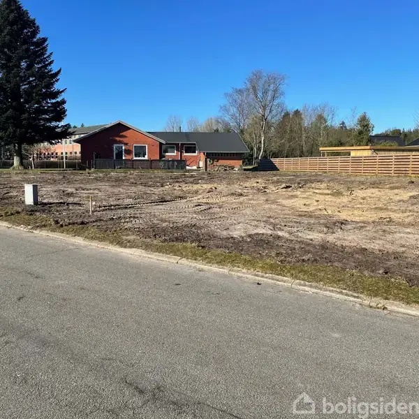Grunden er tom med sparsom vegetation, med et rødt murstenshus i baggrunden. Der er en bred vej i forgrunden, og himlen er klar og blå.
