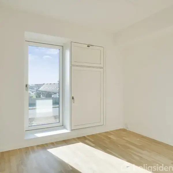 Et tomt værelse med trægulv, en radiator og lysindfald fra et stort vindue. En indbygget skabsdør ses på væggen. Uden for vinduet er der udsigt til tage.