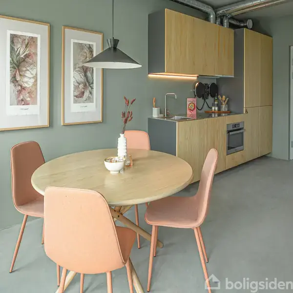 Rundt bord med lysebrune stole, dekoreret med vase og skål, placeret foran et moderne køkken med trækabinetter. Grønlige vægge prydet med to botaniske malerier skaber en rolig atmosfære.