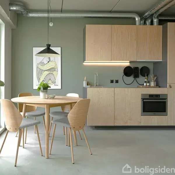 Et rundt spisebord med fire stole står i et lyst køkken. Køkkenskabe i træ hænger på væggen, og en plante er placeret ved et stort vindue.