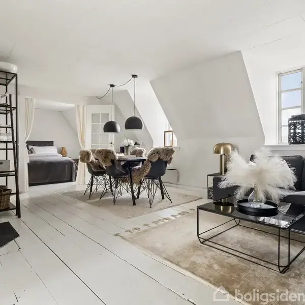 Stue med grå sofa, sort sofabord, og spisebord med stole. Brændeovn til venstre, og soveområde i baggrunden. Rummet er lyst med hvide vægge og gulve.