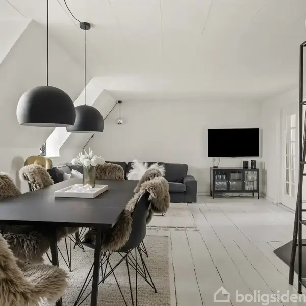 Et moderne spisestueområde med sorte lamper over et spisebord. Stolene har pelsbetræk. I baggrunden ses en stue med sofa, tv og hylder. Gulvet er hvidt træ.