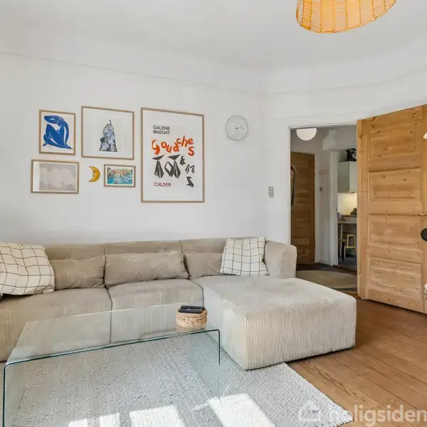 Beige hjørnesofa står i en lys stue med trægulv og glasbord. Væggen er dekoreret med forskellige kunstværker og plakater. En trædør fører til et tilstødende rum.