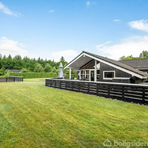 Et gråmalet hus står stille i et åbent, græsbeklædt område omkranset af lave hegn. I baggrunden ses en tæt række af høje træer under en blå himmel.