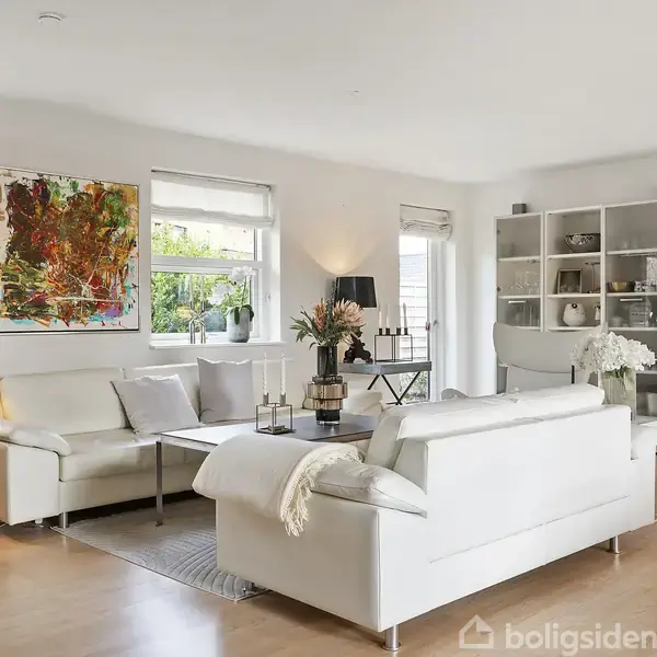 Stue med hvide sofaer står omkring et sofabord med blomster. Et abstrakt maleri hænger på væggen, og et vitrineskab fyldt med vaser er i baggrunden.