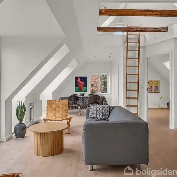 En moderne stue med en grå sofa og et rundt træbord. Stigen fører til en hems. Store vinduer giver naturligt lys. Plakater pryder væggene.