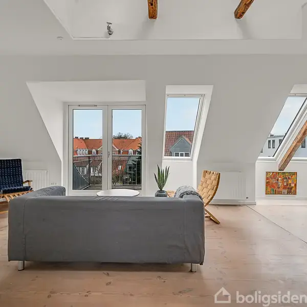 En grå sofa står i en stue med træbjælker i loftet. Store vinduer bagved giver udsigt til røde hustage. Rummet indeholder også stole, et bord og kunst på væggene.