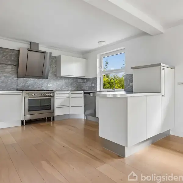 Moderne køkken med hvide skabe og stålovn. Lys træbund, grå flisebagvæg. Store vinduer giver udsigt til grønne omgivelser. Hvidt loft og vægge skaber en lys og åben atmosfære.