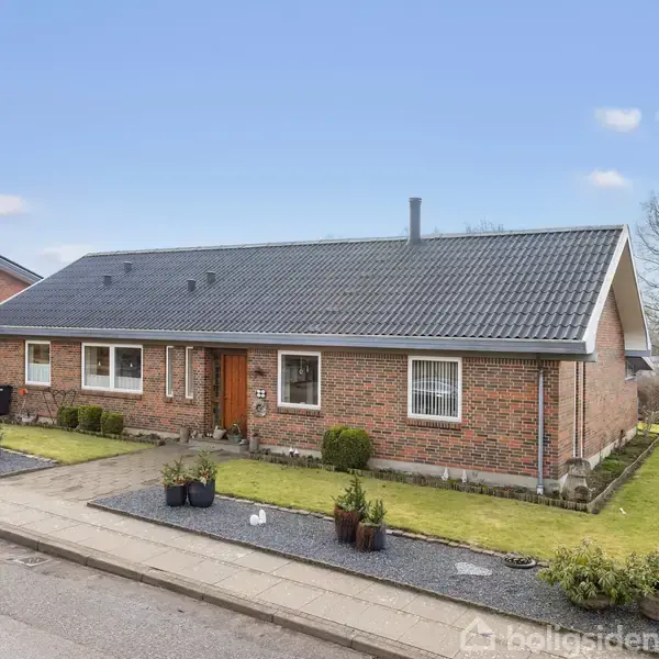 En murstensvilla med sort tag står til salg, omgivet af velplejet have og flisebelagt gangsti. Skyfri blå himmel i baggrunden. Skiltet "Til Salg" ses foran huset.
