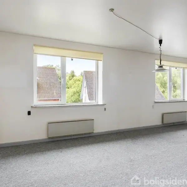 Et tomt rum med hvidmalede vægge og grå gulvtæppe. To store vinduer slipper lys ind og tilbyder udsigt til træer. En enkel loftslampe hænger fra loftet.