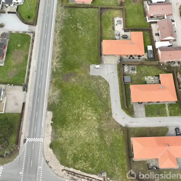 Vejen strækker sig gennem et boligområde, omgivet af huse med røde tage. Græsarealer adskiller ejendommene, og kun få biler er synlige på vejen.